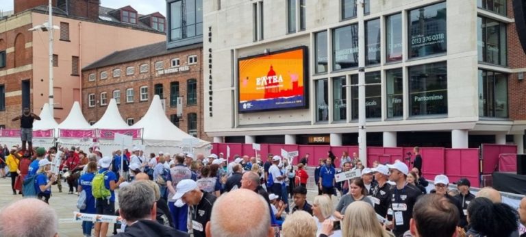 A large group of people, some wearing hats and white shirts, gather in a city square for an event, with a digital billboard displaying an advertisement in the background.