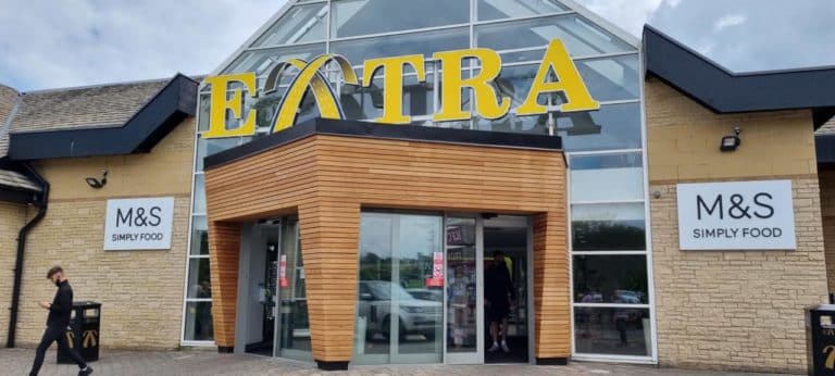 The entrance of a motorway service area with "extra" signage above, flanked by M&S Simply Food stores on either side, and a person walking past.