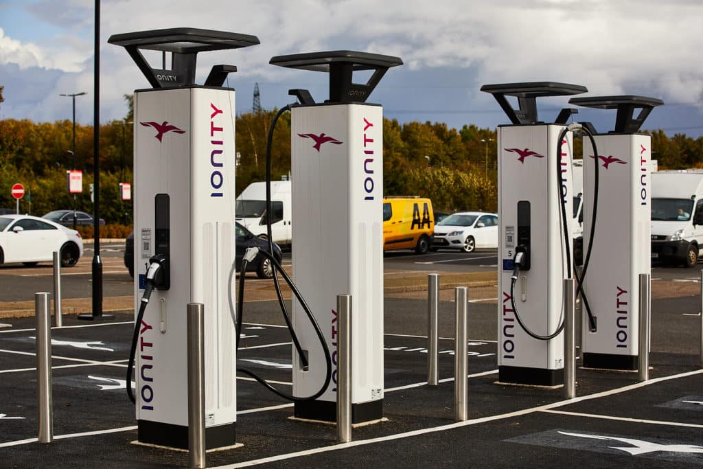 Electric vehicle charging stations and food outlets in a parking lot under a cloudy sky.