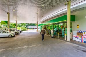 A person walking towards the entrance of a gas station convenience store, with cars parked at fuel pumps under a covered forecourt.