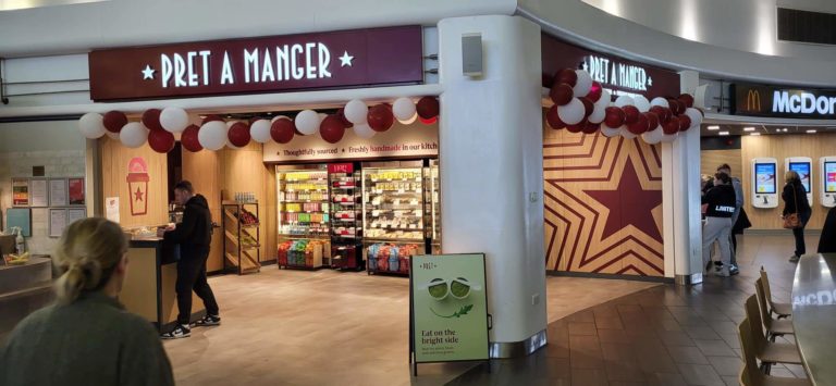 Pret a manger store with red and white balloons decorating the entrance, located next to a mcdonald's in a bustling indoor setting.