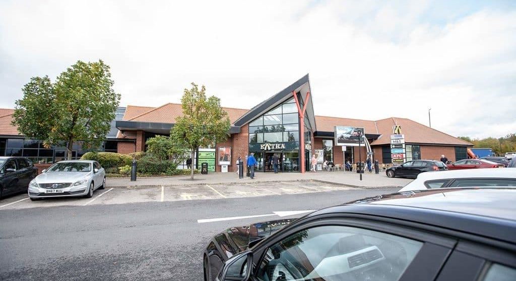 Modern shopping center with a distinctive triangular entrance, under a cloudy sky, with vehicles parked in the foreground near the motorway.