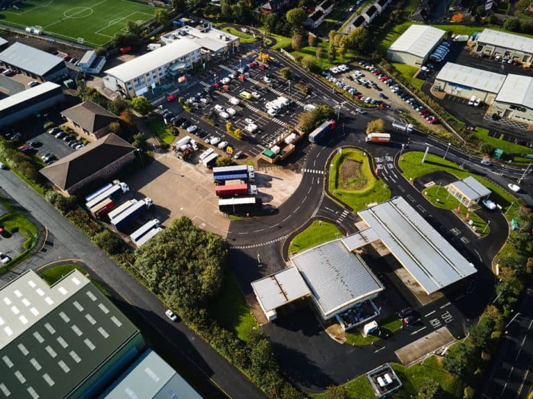 Aerial view of an industrial area with buildings, parking lots, and trucks on motorways, surrounded by green patches.