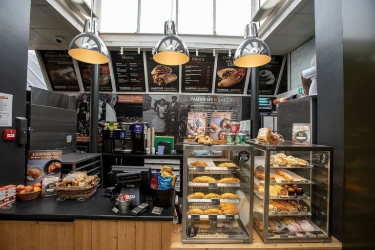 Interior of a Motorway Service Area bakery with a display of assorted pastries and breads, under menu boards with prices, lit by hanging lamps.