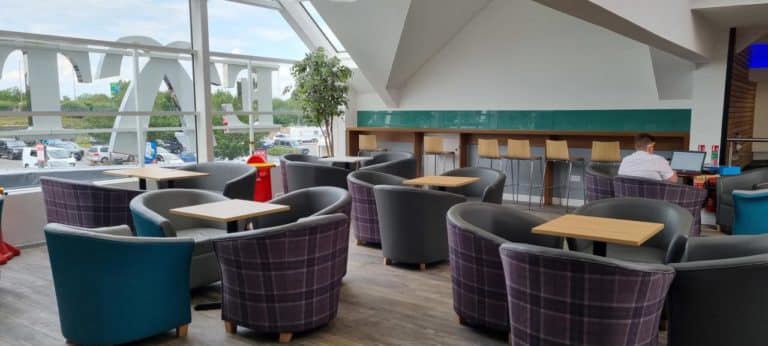 Modern cafeteria located in a motorway service area, with purple and blue chairs, wooden tables, and large windows overlooking a parking area.