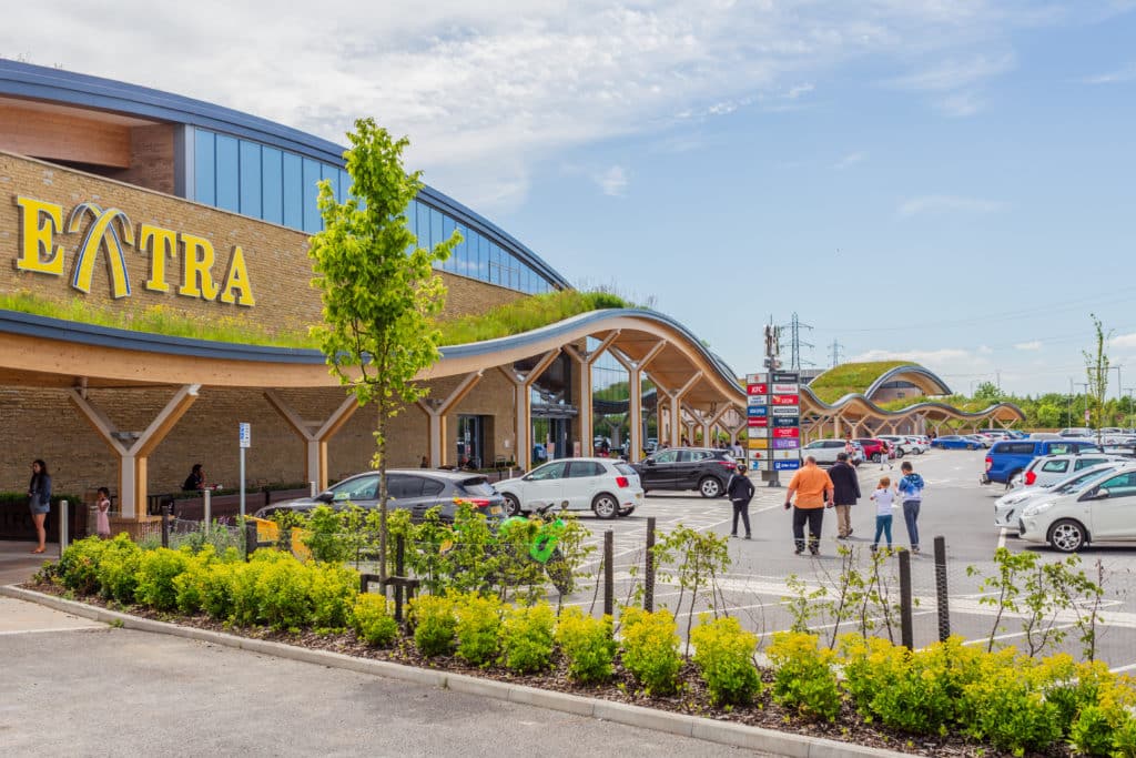 Exterior view of a modern extra supermarket with arched wooden canopy, featuring parked cars and shoppers on a sunny day.